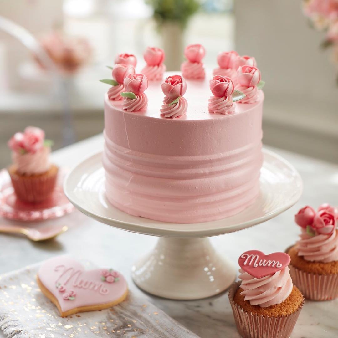 A cake covered in pink frosting with small candy roses assembled in a circle on top. Cookies and cupcakes around are labeled "Mum."