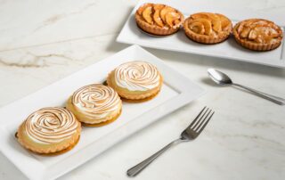 Two plates of three tarts each. One plate's tarts are filled with apple slices, and the other's have swirls of cream on top.