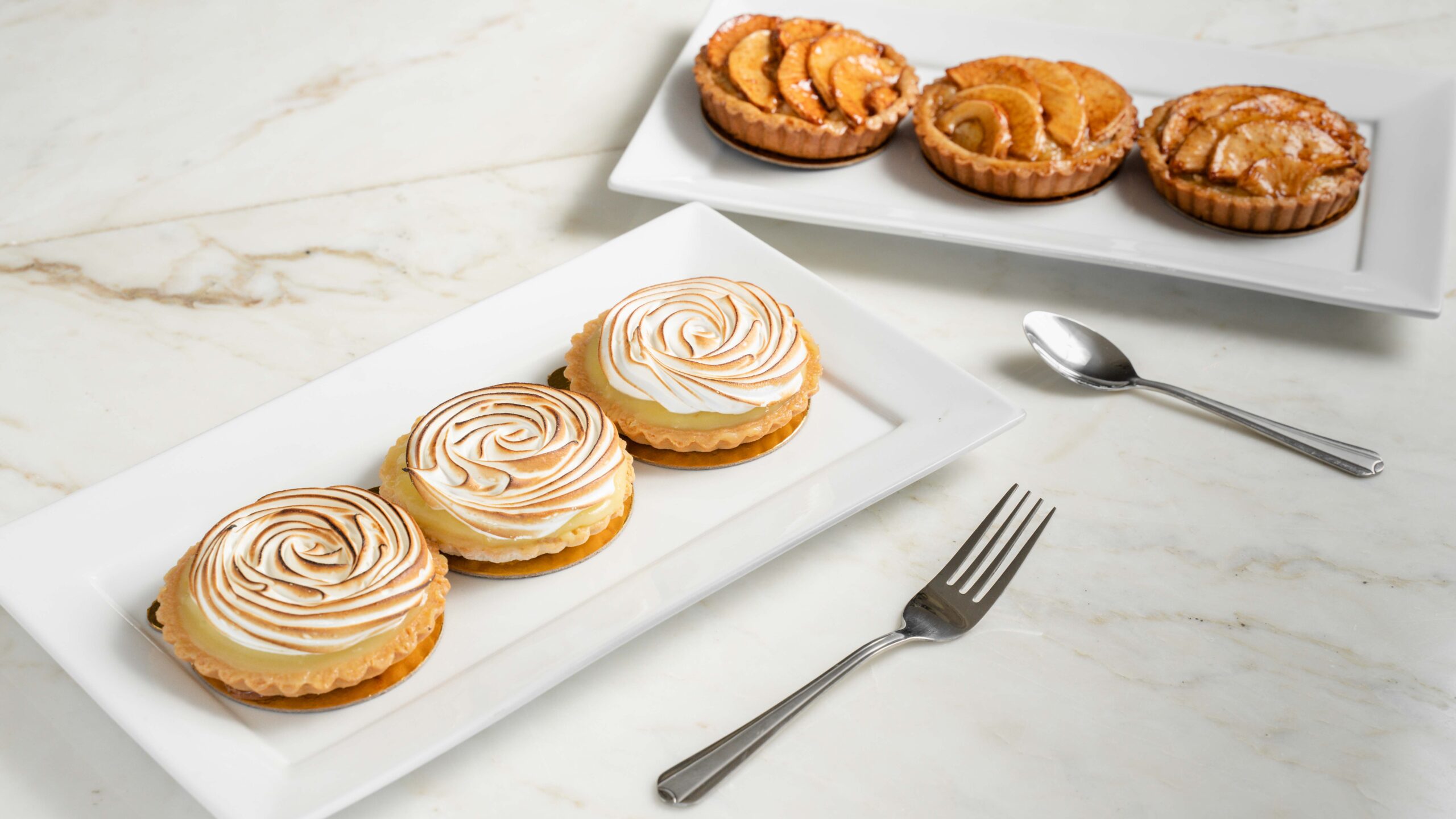 Two plates of three tarts each. One plate's tarts are filled with apple slices, and the other's have swirls of cream on top.