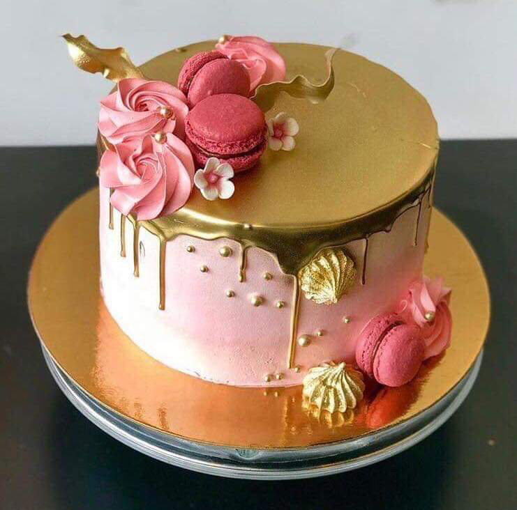 A cake covered in pink frosting with gold icing dripping down from the top. It's embellished with pink macarons and swirls of pink and gold frosting.