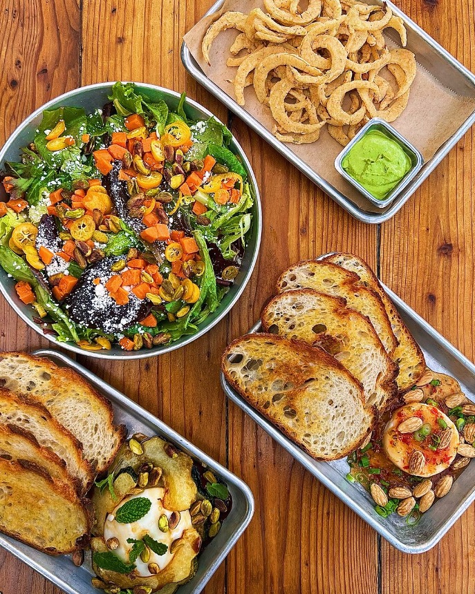 A salad, a plate of onion rings, and two trays with bread slices and different dips.