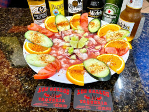 A plate of ceviche with slices of orange, tomato, and cucumber, surrounded by beer bottles.