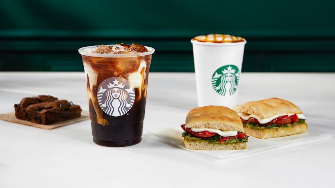 A cold drink and a warm one, both in Starbucks cups, alongside two sandwiches and a brownie.